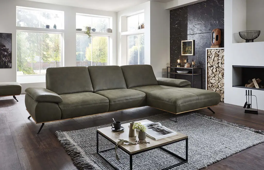 Großes Ecksofa in Olivgrün mit Lederoptik, schwarzen Metallfüßen und gemütlichen Armlehnen. Modernes Wohnzimmer mit Kamin, Holzdeko und großem Teppich – perfekte Kombination aus Komfort und Design.